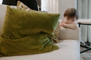 Young asian toddler with her face in her arm looking off while leaning on a white couch with green throw cushion