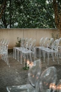  Set up of a wedding ceremony at Marie Gabrielle with clear acrylic ghost chairs within a grove of trees with string lights hanging and more lights wrapped around the base of each tree