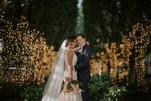  Bride and groom holding each other and smiling at the camera in a grove of lighted trees behind them