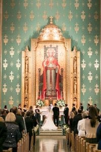  Bride groom and their bridal party standing at the altar in front of the priest as guests look on A large image of Christ is in the background