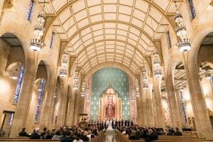 Bride groom and their bridal party standing at the altar in front of the priest as guests look on A large image of Christ is in the background