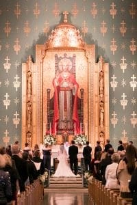 Bride groom and their bridal party standing at the altar in front of the priest as guests look on A large image of Christ is in the background