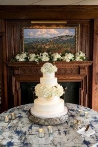 A white four tier wedding cake decorated with hydrangea and roses with a monogram on the second tier from the bottom