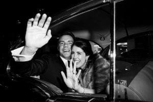 Bride and groom waving laughing and waving goodbye to guests as they are in their getaway car ready to drive off