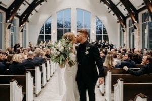 Bride and groom kissing as they walk out the chapel affer their ceremony and bride is holding her bouquet as guests look on