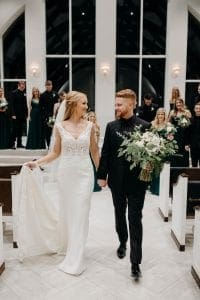 Bride and groom smiling at each other as they exit the wedding ceremony chapel as the groom holds the bridal bouquet and the bride holds her wedding train and their bridal party looks on