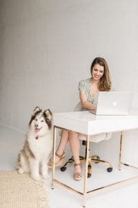 Smiling woman sitting on an ivory rolling office chair typing on a macbook air on a sleek white desk while a brown and white Siberian Husky sits by her side