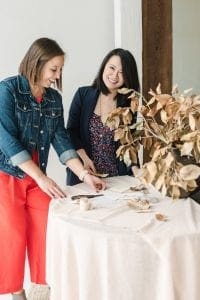 Two smiling women arranging a stationery suite hovering over a table with blush linen