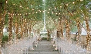 Set up of a wedding ceremony at Marie Gabrielle with clear acrylic ghost chairs within a grove of trees with string lights hanging and more lights wrapped around the base of each tree with a floral arch in the front