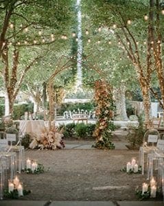 Set up of a wedding ceremony at Marie Gabrielle with clear acrylic ghost chairs within a grove of trees with string lights hanging and more lights wrapped around the base of each tree with a floral arch in the front