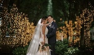 Bride and groom holding each other and smiling at the camera in a grove of lighted trees behind them