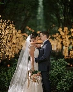 Bride and groom holding each other and smiling at the camera in a grove of lighted trees behind them