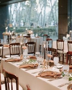 A wedding reception setup at Marie Gabrielle with white linens and brown bistro chairs There are brown charger plates and rust napkins on the table settings with glassware