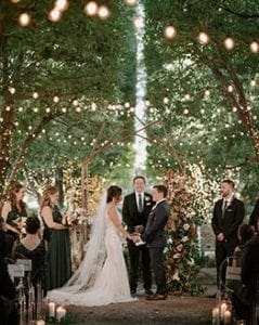  Bride and groom standing at the altar with their bridal party and officiant under a grove of lit trees and string lights attached from one end to another in front of a metal and floral arch