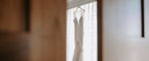  White wedding gown hanging on a hanger by a window with white drapes