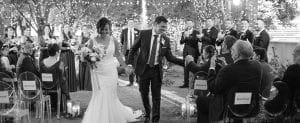 Bride and groom smiling as they exit their wedding ceremony while bridal party and guests look on and the groom shakes the hand of a guest