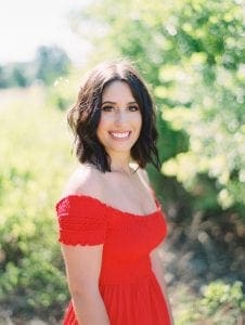 Smiling headshot of a brunette with brown eyes standing in front of a green backdrop wearing an off shoulder red dress