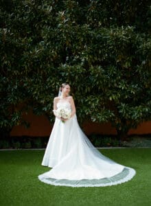 Bride in a white strapless wedding gown with a long train and bouquet standing on a green lawn in front of dense hedges smiling