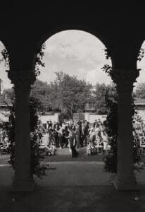 Outdoor wedding ceremony under stone arches guests mingling beside decorated chairs in a garden setting black and white
