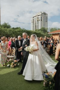 Bride in a white wedding gown and veil walks beside an older man in a tuxedo holding a bouquet wedding guests smile and watch outdoors