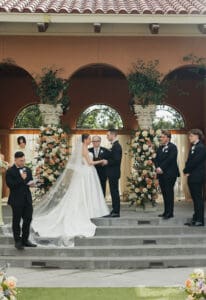 Bride and groom exchange vows on stone steps officiant reading with wedding party in black tuxedos beside rose filled arches