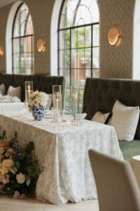 Elegant event table set with a white patterned tablecloth tall glass candle holders and a blue and white vase of white flowers against a tufted green banquette by arched windows