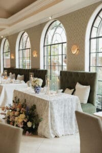 Elegant dining table setup with a white lace tablecloth floral centerpiece and tall candles against arched windows and tufted seating