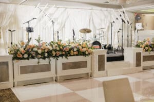 Wedding reception stage with white draped backdrop and long floral garlands along the front of a raised white dais microphone stands and a drum kit in the background