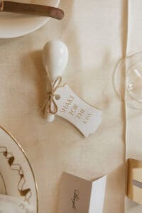White ceramic spoon like decor tied with twine and a tag that says Shake for the kiss on a beige table setting with plates and glassware nearby