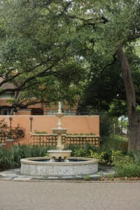 Stone tiered fountain in a courtyard garden with lush greenery and a peach colored building in the background