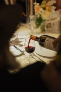 Red wine being poured from a bottle into a stem glass at a formal dinner table with a blurred floral centerpiece in the background