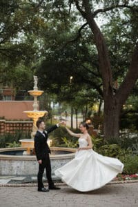 Bride in a white strapless gown spins as groom in a black suit holds her hand beside a decorative fountain in a garden setting