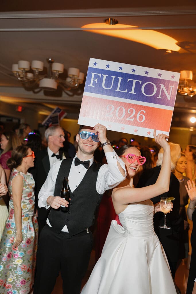 Bride and groom at their wedding reception cheerfully holding a celebratory sign amid dancing guests