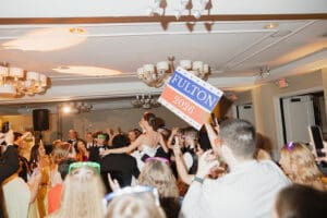 A person hoists a Fulton 2026 sign above a crowded party with guests dancing and cheering in formal attire