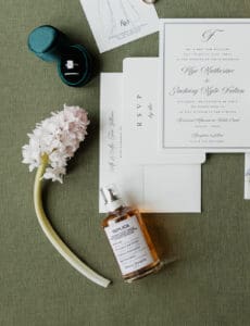 Flat lay of wedding details invitations with calligraphy a diamond ring in a teal ring box a white hyacinth and a Replica perfume bottle on olive fabric