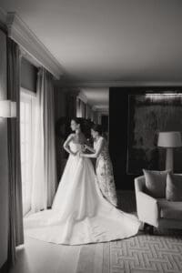 Bride in a strapless wedding gown being helped with the back by a woman in a hotel room