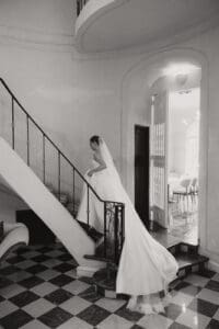 Bride in a long wedding dress and veil descends a curved staircase in a grand black and white foyer