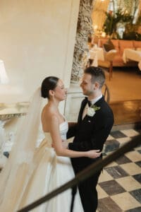 Bride in a white gown with veil facing a groom in a black tuxedo both smiling and embracing at a wedding venue