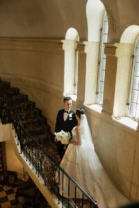 Bride in a white gown with veil and bouquet talks with a groom in a black tux on a grand staircase in a historic interior lit by large arched windows