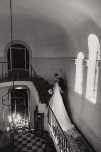 Bride in a wedding gown with veil and bouquet on a curved staircase beside a groom in a dark suit inside a grand historic interior