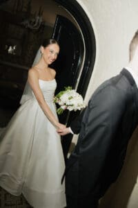Bride in a strapless white wedding gown and veil smiles as she holds a bouquet of white flowers entering through a large arched doorway while holding hands with a groom in a black suit