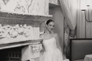 Bride in a strapless wedding gown leaning beside an ornate stone fireplace in a vintage room