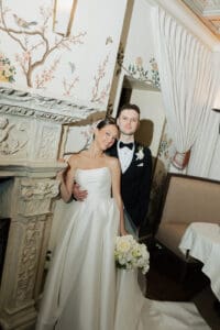 Bride in a strapless white wedding gown holds a white bouquet as she leans beside a carved stone mantel with pink floral wallpaper and a smiling groom in a black tuxedo beside her in an elegant room shift