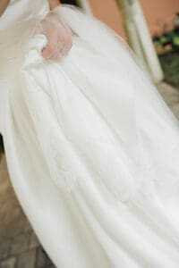 Close up of a brides white wedding dress with lace hem held by a hand with a ring veil softly draped over the fabric