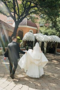 Bride in a white gown and veil walks beside a groom in a black suit through a sunlit courtyard with white canopy tents in the background