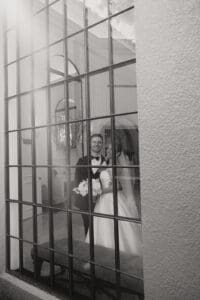 Bride and groom in wedding attire smile together viewed through a large grid window bouquet in hand