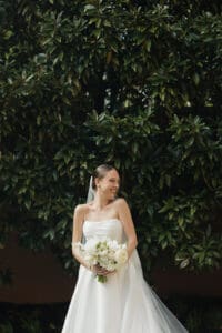 Bride in a strapless white wedding gown smiling holding a bouquet of white roses outdoors in front of a dense green hedge
