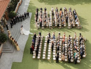 Aerial view of an outdoor wedding with a bride in a white gown approaching the ceremony area lined with rows of beige chairs and floral arrangements on a grassy lawn