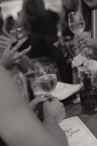 Close up of a person holding a wine glass at a table with a small flower vase and a menu nearby others blurred in the background