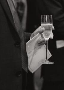 Person in a suit holds a champagne flute and a folded thank you card at a formal event ready to toast